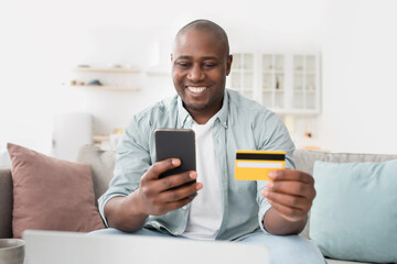 Fast online shopping. Cheerful african american man with credit card and smartphone sitting on couch at home