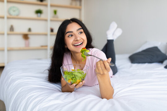 Weight loss diet concept. Pretty Indian woman eating fresh vegetable salad on bed at home, full length