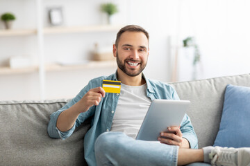 Happy young guy with credit card and tablet computer sitting on couch at home, purchasing goods on web