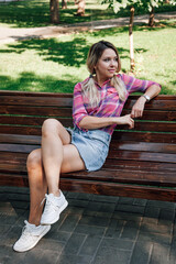 Adult blonde woman with face looking away sitting on bench in park with left wrist on backrest...