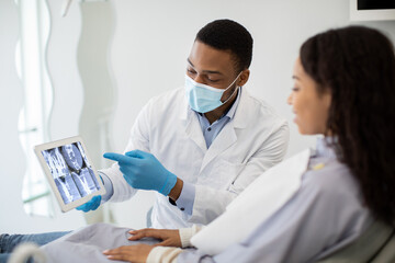 Black Dentist Doctor Showing Treatment Result On Digital Tablet To Female Patient