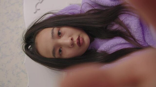 Vertical video mobile phone POV of 20s Asian woman typing message lying in bed. Woman in purple pajamas using cell phone.