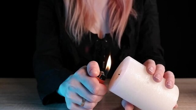 Woman lights a white rustic candle with a lighter over a wooden table in super slow motion. Sparks and flame are visible against a girl in a black suit with long hair who sits on a black background