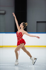 side view of happy woman in red dress figure skating with outstretched hands in professional ice rink