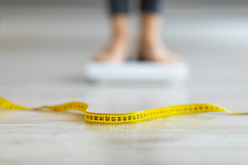 Measuring tape on floor and blurred view of millennial woman on scales, checking result of her slimming diet