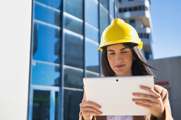 Young beautiful woman in brown coat, yellow workman's helmet and blueprint tube, working with her tablet. Business concept, architect, construction, working woman, empowerment.