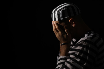 Remorseful prisoner in striped uniform with handcuffs hiding his face on black background, space...