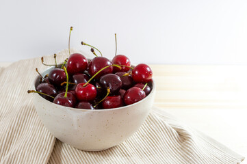 Organic red cherries in a ceramic bowl and a napkin on a light wooden table, healthy fruits as summer snack, copy space, selected focus