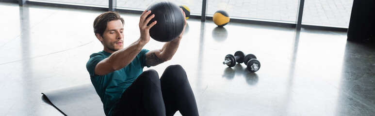 Brunette sportsman working out with slam ball near dumbbells in gym, banner