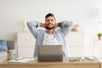 Taking break. Happy freelancer guy leaning back in chair, resting at workplace with hands behind...