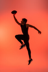 Fototapeta na wymiar Side view of athletic player holding rugby ball while jumping isolated on red