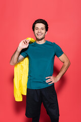 Positive sportsman holding yellow sports jacket and hand on hip on red background