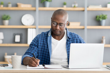Fototapeta na wymiar Concentrated black mature man attending online training from home in front of laptop and taking notes