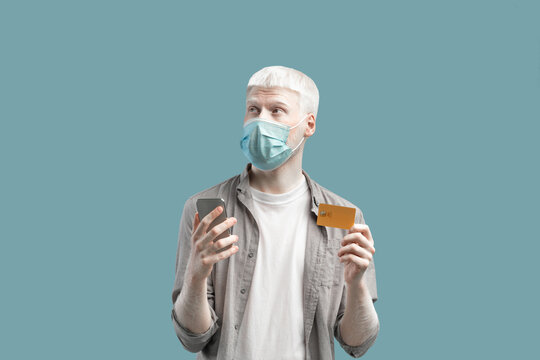 Thoughtful albino guy in face mask using credit card and smartphone to shop online on turquoise background. - Powered by Adobe