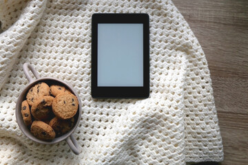 Digital e-reader or tablet, bowl of chocolate chip cookies and soft knitted blanket on wooden...