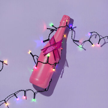 Christmas creative layout with pink champagne bottle and colorful christmas lights string on pastel purple background. 80s or 90s retro fashion aesthetic party concept. New Year celebration idea.