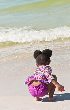 Back View, Close Distance Of A Black Female Child, In A Purple Bathing Suit At A Tropical Shoreline Collecting Sea Shells 