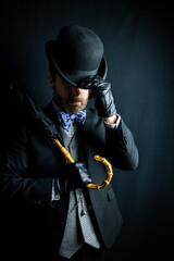 Fototapeta premium Portrait of British Gentleman in Dark Suit Holding Umbrella Over His Shoulder and Doffing Bowler Hat. Vintage Style and Retro Fashion of Film Noir Secret Agent Spy Hero.