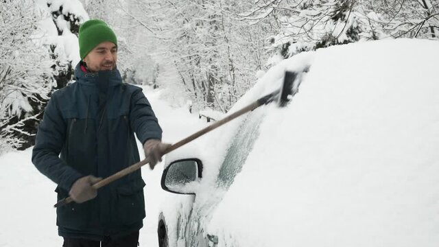 Man cleaning snow-covered car with brush after heavy snowfall. Snowstorm, bad winter weather, natural disaster. Person cleans side windows of his car from snow.
