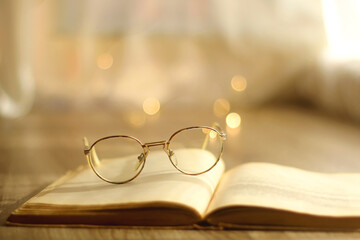 Open book and reading glasses on the table. Bokeh lights in the background. Hygge at home. Selective focus.