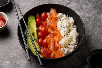 Poke bowl - black bowl with white rice, smoked salmon, cherry tomatoes and avocado, sesame seeds, soy sauce and chopsticks on a grey concrete surface, close-up
