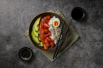 Poke bowl - green bowl with white rice, smoked salmon, cherry tomatoes and avocado, sesame seeds, soy sauce and chopsticks on a grey concrete surface, top view