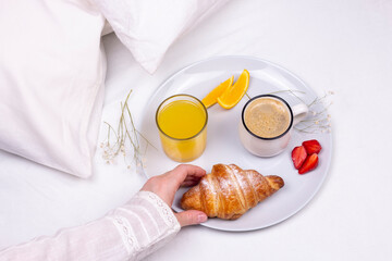 woman's hand takes a croissant, romantic surprise breakfast in the bed. Croissant, cup of coffee, orange juice on the ceramic plate. Sweet morning. flat lay top view image.