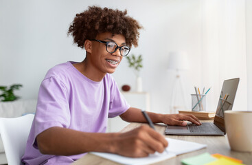 Home schooling concept. Smart black teen guy using laptop at table, writing in notebook during...