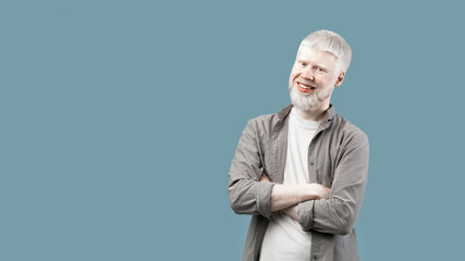 Positive albino guy with folded arms looking and smiling at camera, turquoise background with empty...
