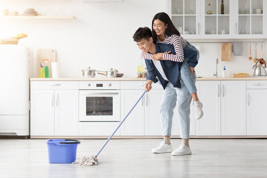 Cheerful asian guy piggyback his pretty girlfriend while mopping kitchen - Powered by Adobe