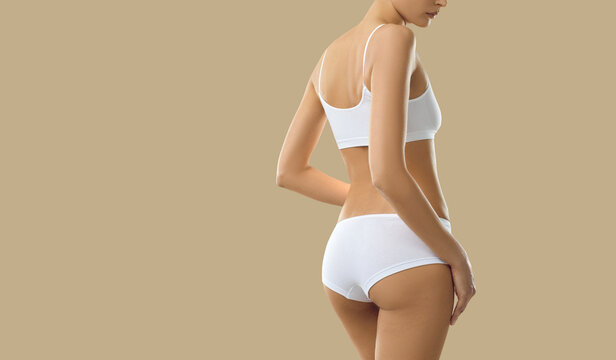 Young model posing in comfy white undies. Woman with beautiful body wearing basic underwear set consisting of soft bra and underpants standing isolated on beige copy space background, view from behind