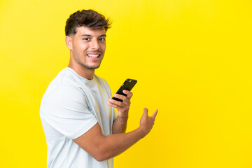 Young caucasian handsome man isolated on yellow background using mobile phone and pointing back