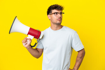 Young caucasian handsome man isolated on yellow background holding a megaphone and thinking