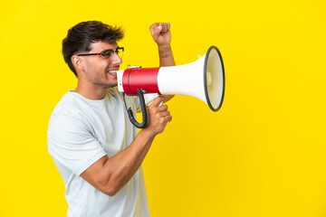Young caucasian handsome man isolated on yellow background shouting through a megaphone to announce something in lateral position