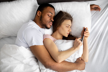 Young woman lying on bed in arms of her black husband and using mobile phone to text her lover, top...