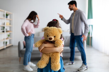 Children and family conflicts concept. Little girl cuddling teddy bear standing against arguing...