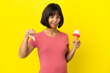 Pregnant woman holding a cornet ice cream isolated on yellow background showing thumb down with negative expression