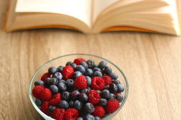 Bowl of blueberries and raspberries and open book on a table. Selective focus.