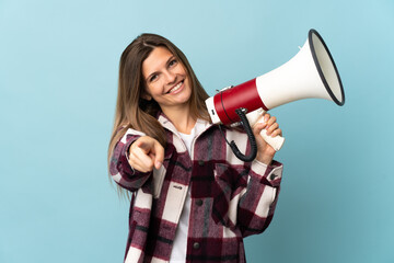 Young Slovak woman isolated on blue background holding a megaphone and smiling while pointing to...