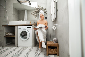 beautiful young woman in a towel sits on the toilet with a magazine 