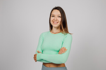 Fototapeta premium Young woman posing in studio. Beautiful brunette in green top for fitness or yoga stands with her arms crossed, smiling at camera on neutral background.