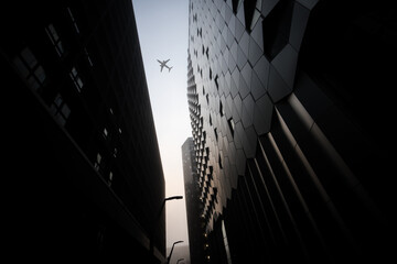Aeroplane jumbo jet passenger plane flying between modern city centre sky scrapers on foggy misty day. Dramatic lighting and steel buildings in futuristic downtown area. - Powered by Adobe