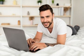 Job in bedroom, browsing on internet. Smiling millennial man woke up, working on laptop, lying on white bed in bedroom