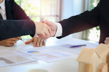Business people shake hands while discussing and sign contract in meeting desk with businessman in black suit blurred background, paper and model house on table, agreement partnership concept.