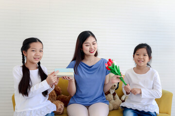 Two daughter girls giving beautiful tulip flowers and present gift box for their happy smiling mother while sitting on sofa in living room. Happy mother day, children have special moment with mom.