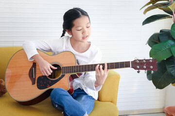 Adorable happy girl singing a song and playing guitar while sitting on sofa in living room, kid...