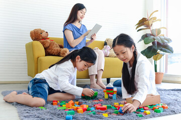 Two daughter girls playing with wooden block game (Jenga) together while mother sitting on sofa and surfing the internet, mom online working by using tablet, sister kid spending time together.