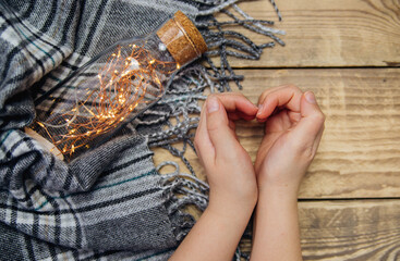 Women's hands make the shape of a heart. A glass bottle with a garland lights .Warm scarf or plaid. A symbol of love.