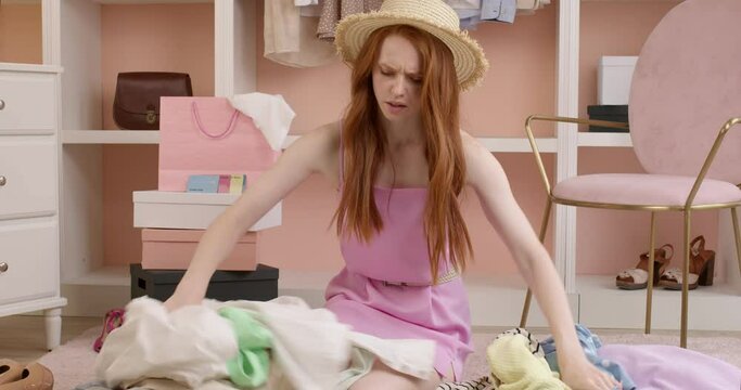 Emotional breakdown. Crying young woman sorting through a pile of clothes from wardrobe, dissatisfied, nothing to wear. redhead upset lady in hat sit on floor feeling frustrated, at a loss, crying