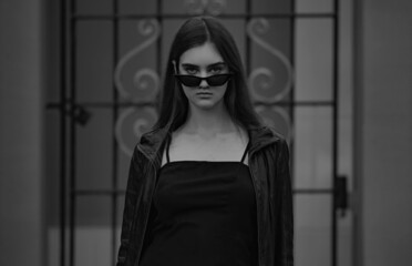 Fashionable style female model in fashion sunglasses posing holding the hands the black leather jacket on blue wall background on the street. Summer and spring style.
